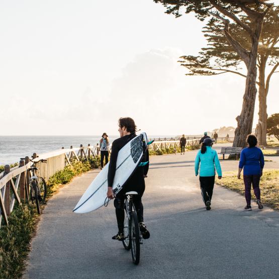 surfer carrying a board and riding a bike on West Cliff 