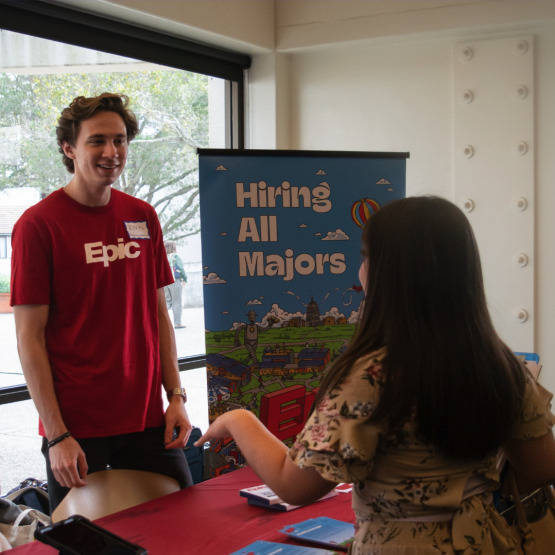 Epic representative  talking to a student behind a table with a banner saying hiring all majors